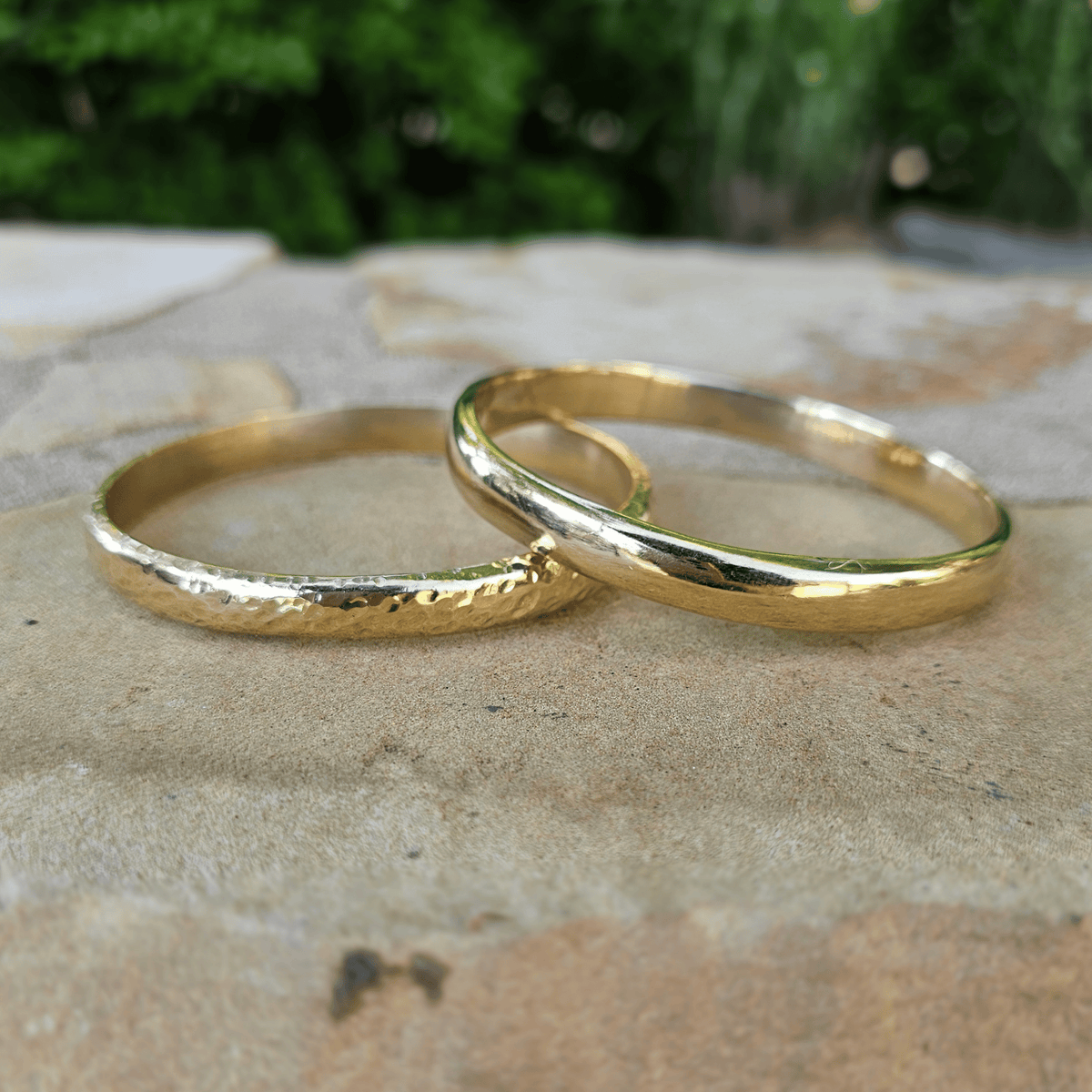 Two gold bracelets, one with a textured hammered finish and one with a smooth polished finish, resting on a stone surface with a blurred green garden background. Shop Kamawe
