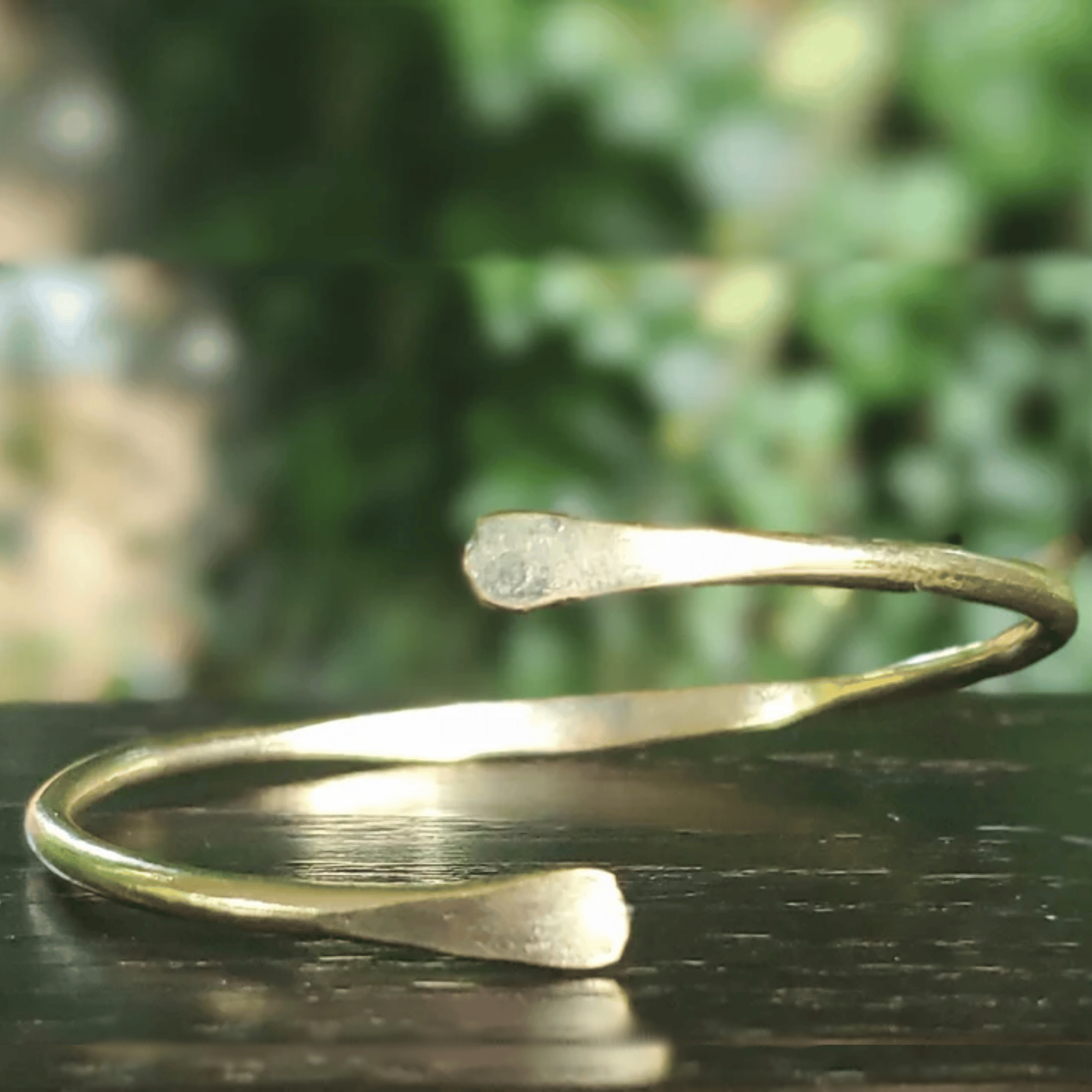 A hammered open cuff bracelet in a warm gold or brass tone, resting on a dark reflective surface with a blurred green outdoor background. Shop Kamawe