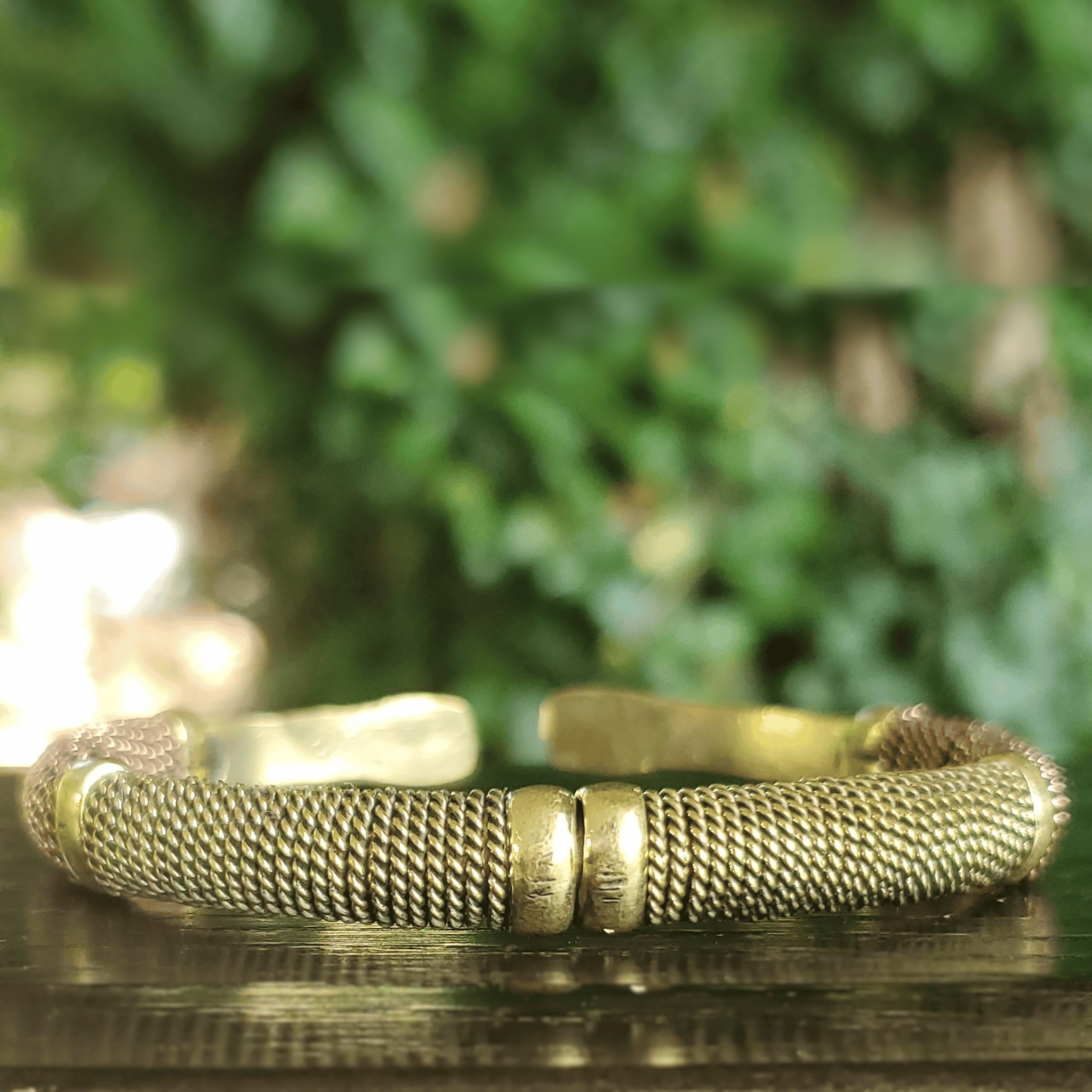 A textured gold-toned metal cuff bracelet with a mesh-like woven pattern and three central cylindrical links, resting on a dark reflective surface with a blurred green foliage background. Shop Kamawe
