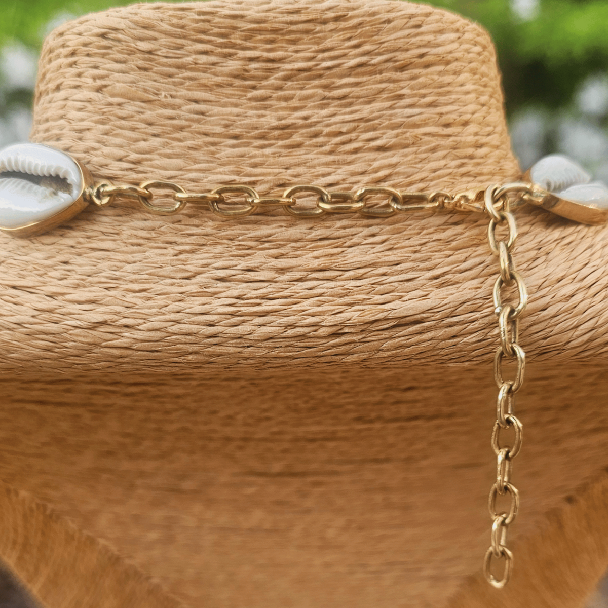 A cowrie shell necklace displayed on a woven beige mannequin bust. The necklace features multiple white cowrie shells with natural brown lines and small gold-toned metal links. Shop Kamawe