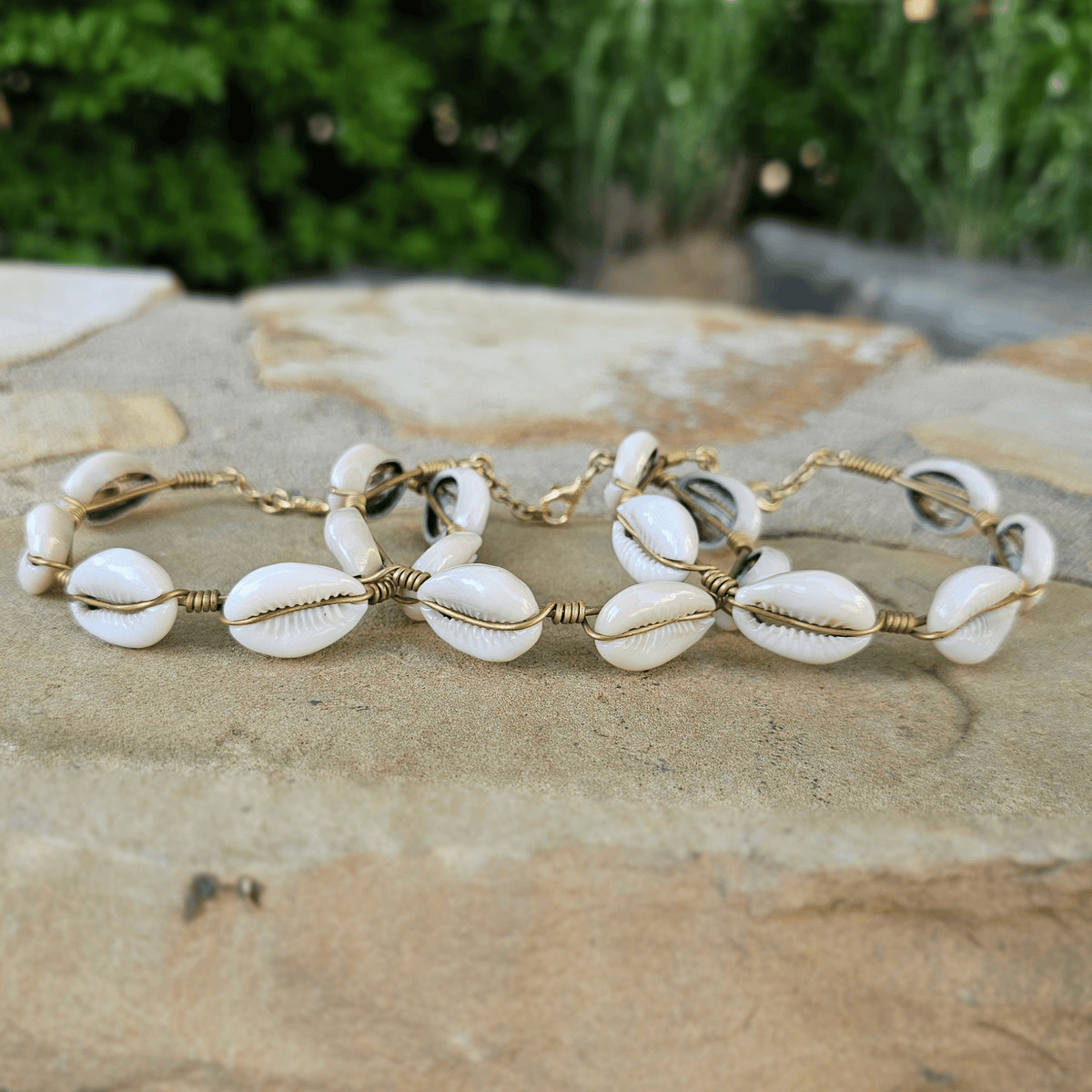 A bracelet made of white cowrie shells and gold wire wrapped details, displayed on a stone surface with a blurred green outdoor background. Shop Kamawe