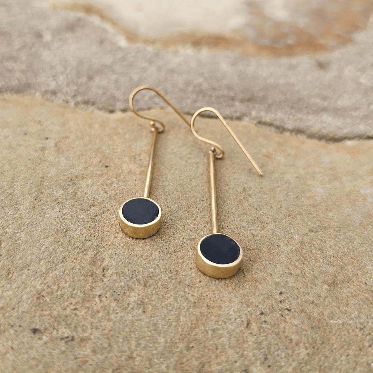 A pair of gold drop earrings with circular disc black pendants, displayed on a textured beige stone surface. Shop Kamawe