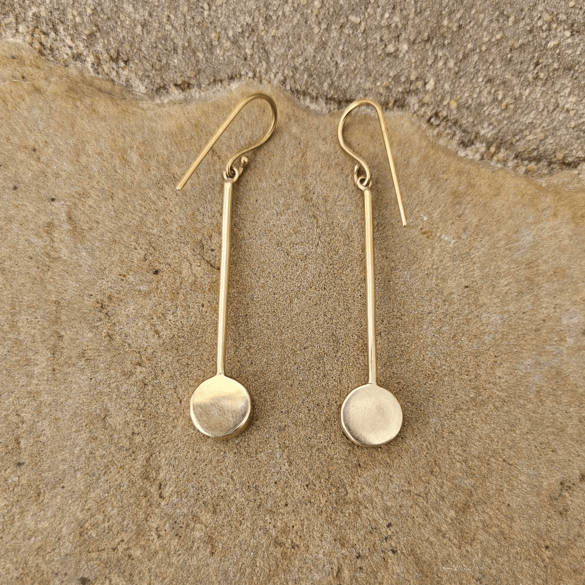  The back side of a pair of gold drop earrings with circular disc black pendants, displayed on a textured beige stone surface. Shop Kamawe