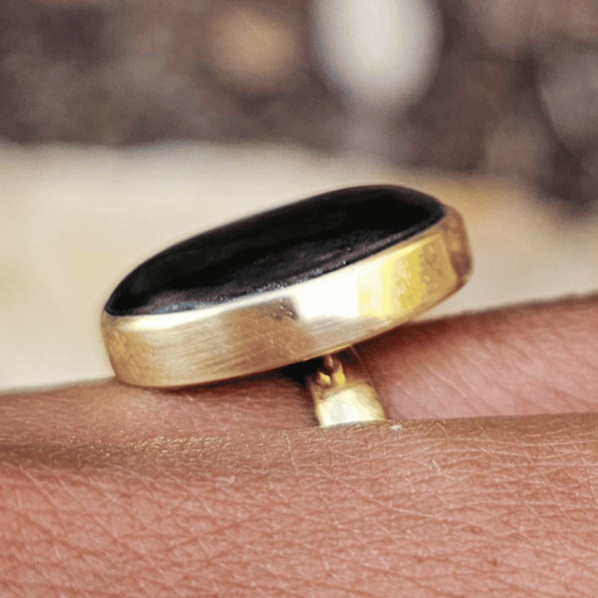 A gold ring with a large oval black cabochon gemstone worn on a person's finger, set against a blurred outdoor background. Shop Kamawe
