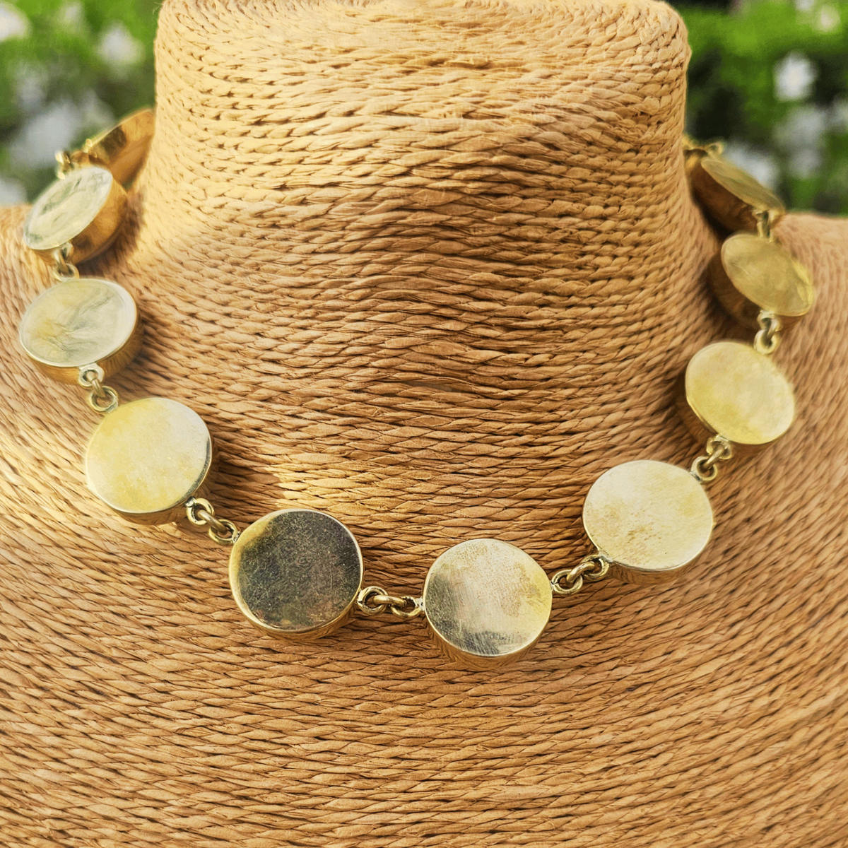 The back side of a gold chain necklace with oval black cabochon stones, displayed on a woven brown mannequin bust with a blurred outdoor background. Shop Kamawe
