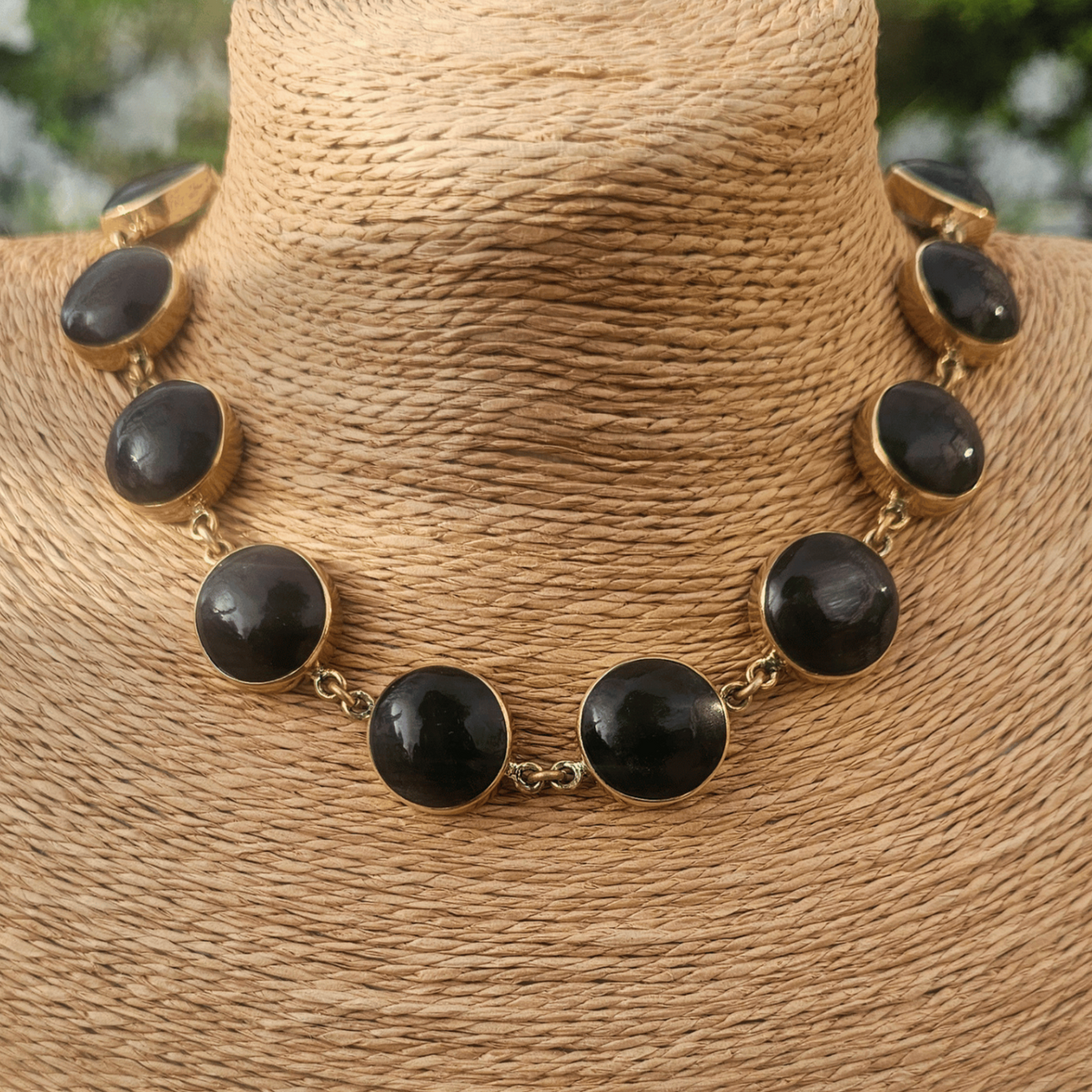 A gold-tone statement necklace with large oval black stones, displayed on a woven tan jewelry bust against a blurred outdoor garden background. Shop Kamawe