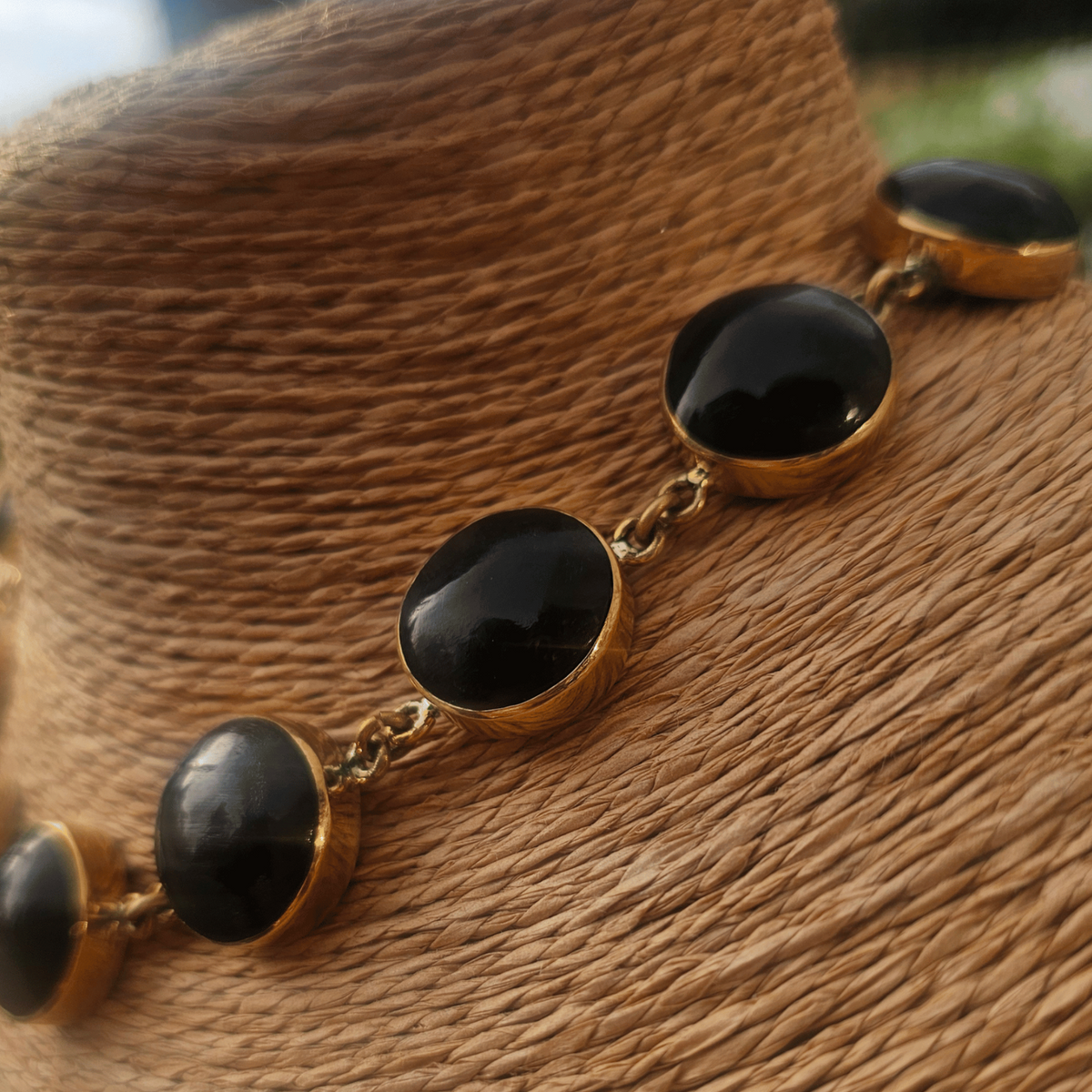 A gold chain necklace with oval black cabochon stones, displayed on a woven brown mannequin bust with a blurred outdoor background. Shop Kamawe