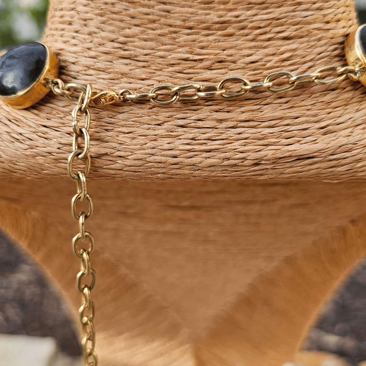 A gold chain necklace with oval black cabochon stones, displayed on a woven brown mannequin bust with a blurred outdoor background. Shop Kamawe