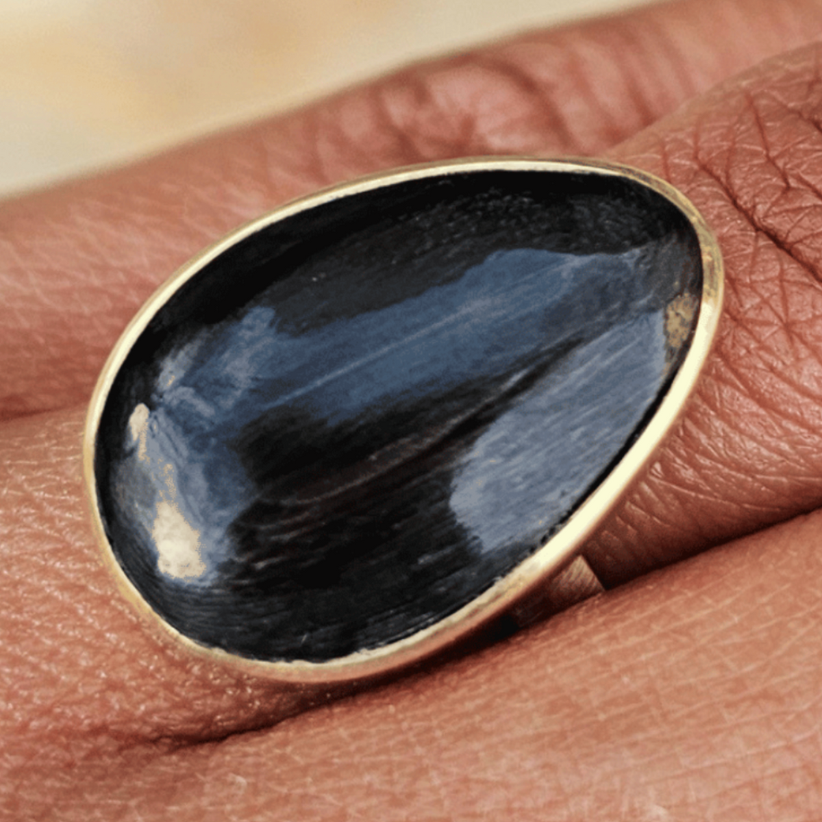 A close-up of a hand wearing a gold ring with a large, smooth, dark teardrop-shaped stone. Shop Kamawe
