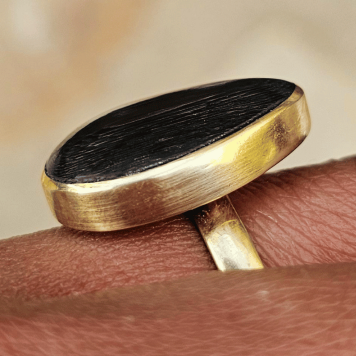 A close-up of a gold ring with a large, polished oval black stone, worn on a person's finger against a soft-focus beige background. Shop kamawe