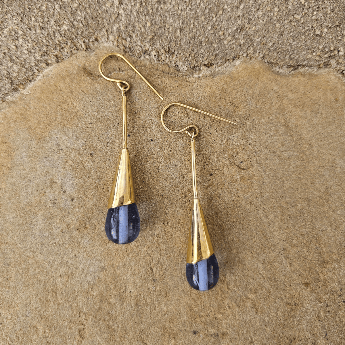 A pair of gold drop earrings with teardrop-shaped blue gemstone pendants, displayed on a textured tan stone surface. Shop Kamawe
