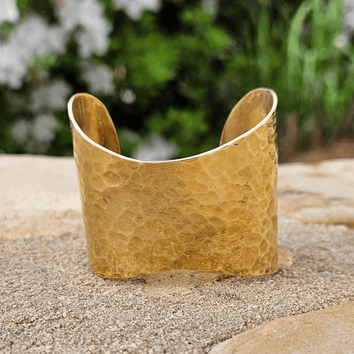 A hammered gold cuff bracelet standing upright on a sandy surface with a blurred green garden background. Shop Kamawe