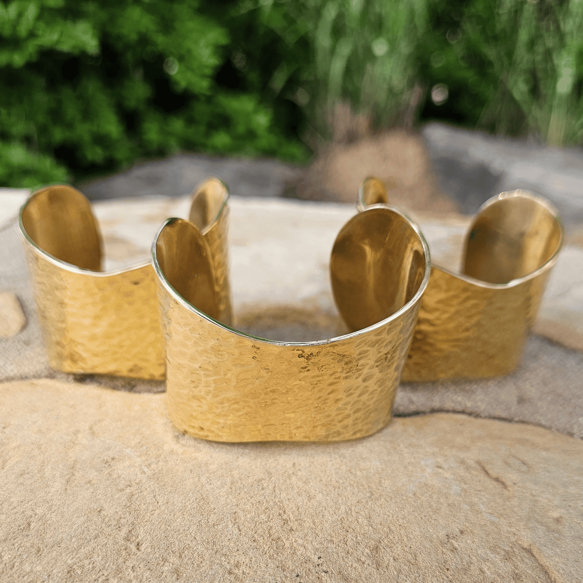Three hammered gold cuff bracelets displayed on a stone surface with a blurred green outdoor background. Shop Kamawe