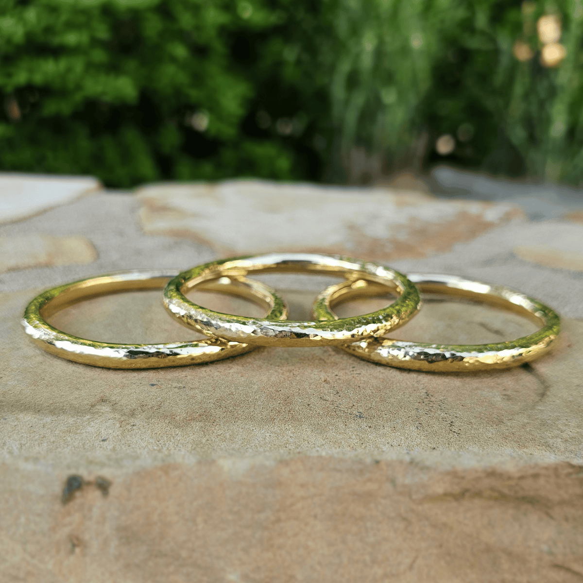 Three textured gold rings arranged overlapping on a stone surface with a blurred green outdoor background. Shop Kamawe