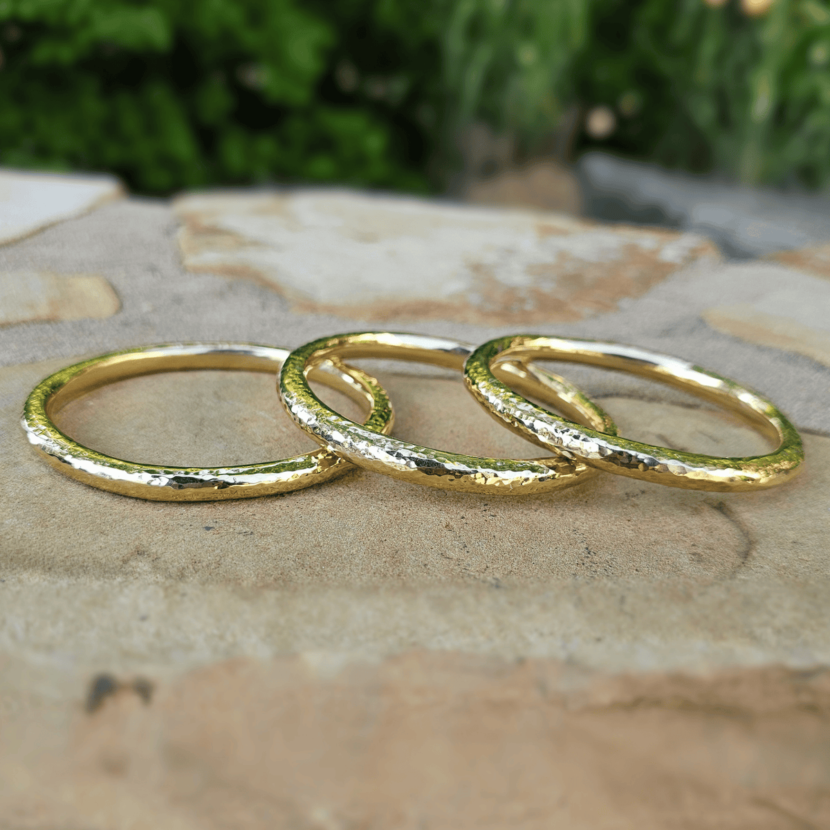 Three textured gold rings arranged overlapping on a stone surface with a blurred green outdoor background. Shop Kamawe