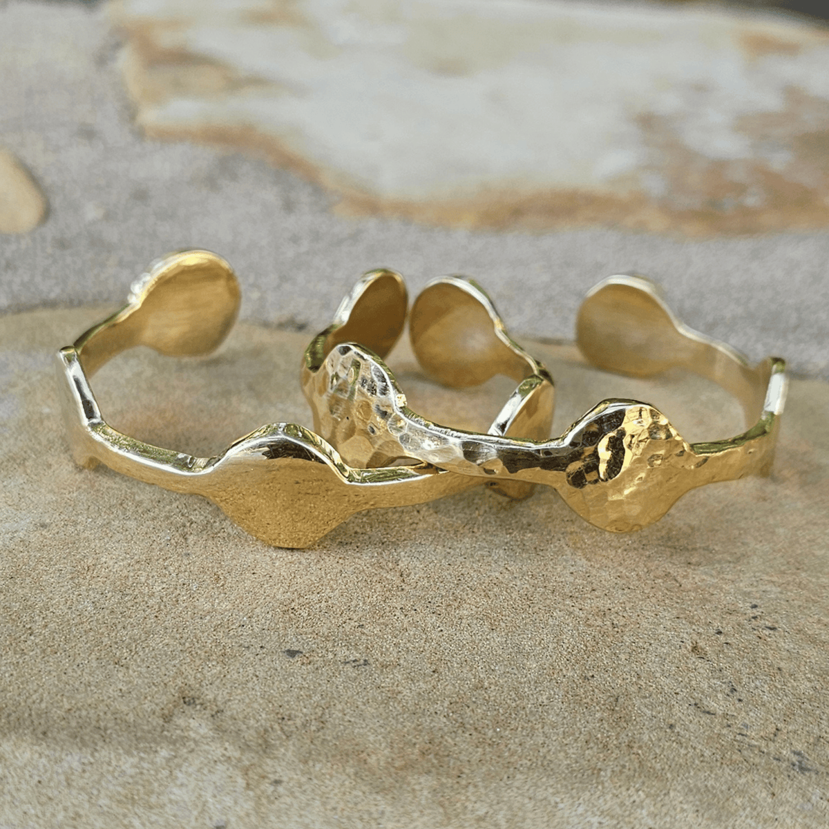 A gold-toned open cuff bracelet with organic, wavy textured shapes, displayed on a neutral stone surface. Shop Kamawe