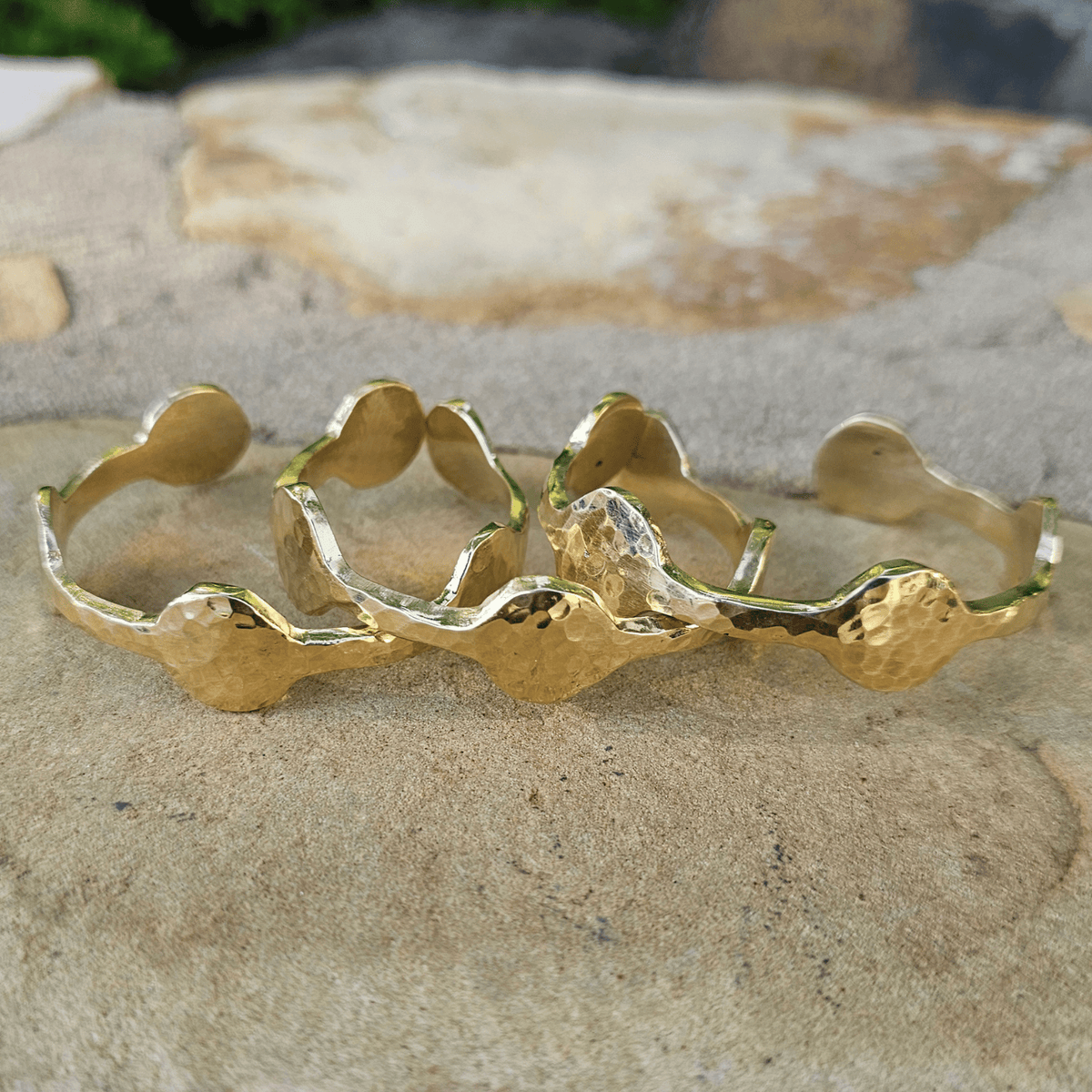 Three hammered gold cuff bracelets with wavy, organic edges, displayed on a textured beige stone surface. Shop Kamawe
