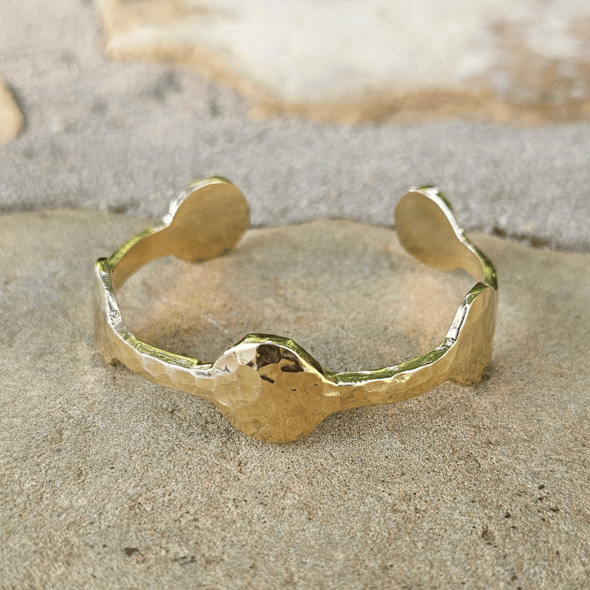 A hammered gold cuff bracelet with organic, wavy edges, resting on a textured stone surface. Shop Kamawe