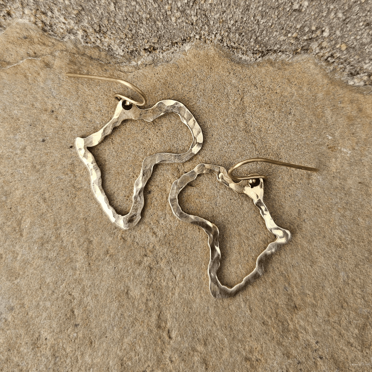 A pair of hammered metal earrings shaped like abstract Africa map landmass, resting on a textured beige stone surface. Shop Kamawe