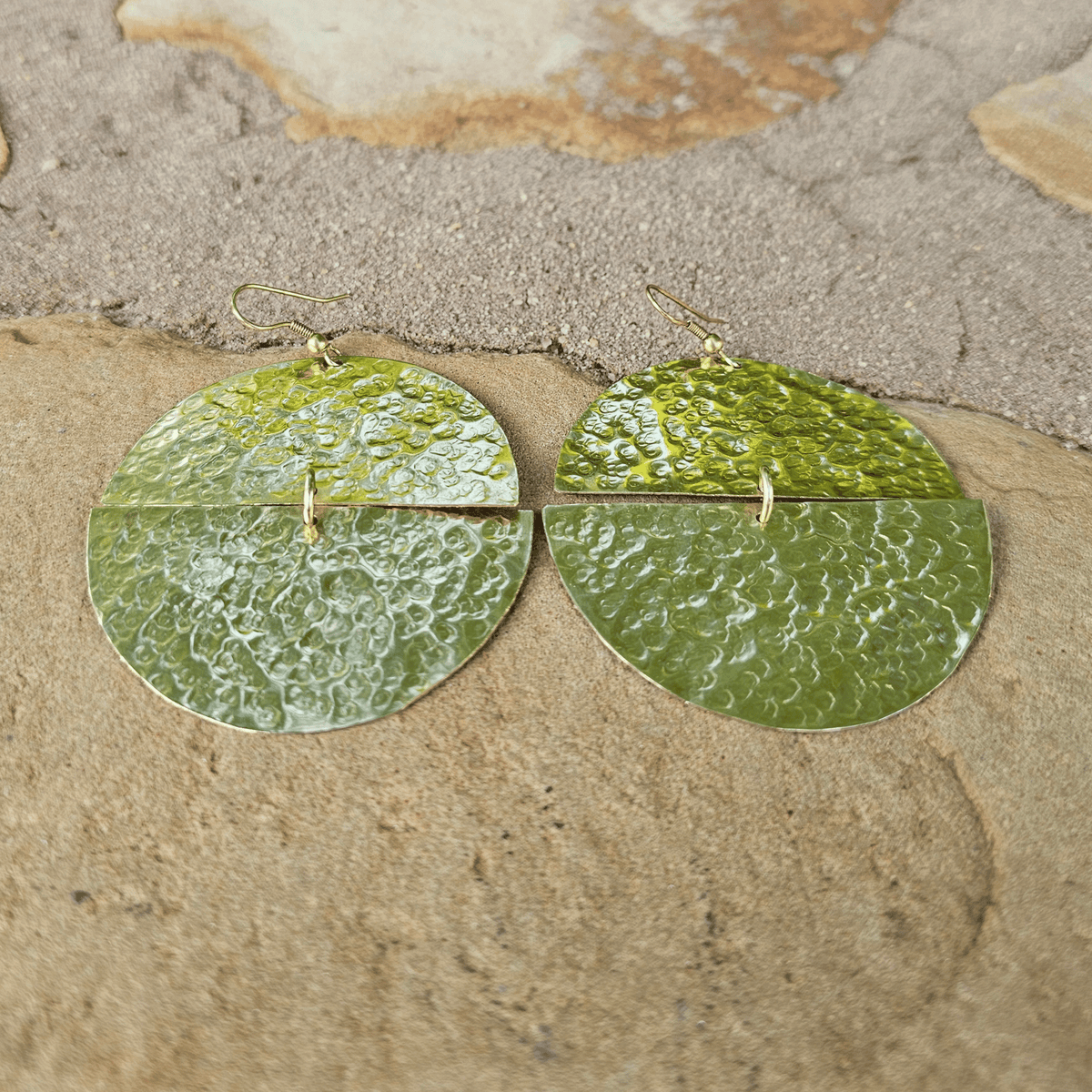 A pair of hammered green metal dangle earrings with a split circular design and small gold hooks, displayed on a textured beige stone surface. Shop Kamawe