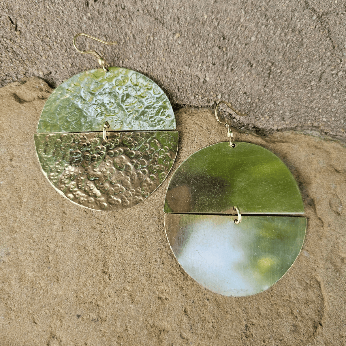 A pair of hammered green metal dangle earrings with a split circular design and small gold hooks, displayed on a textured beige stone surface. Shop Kamawe