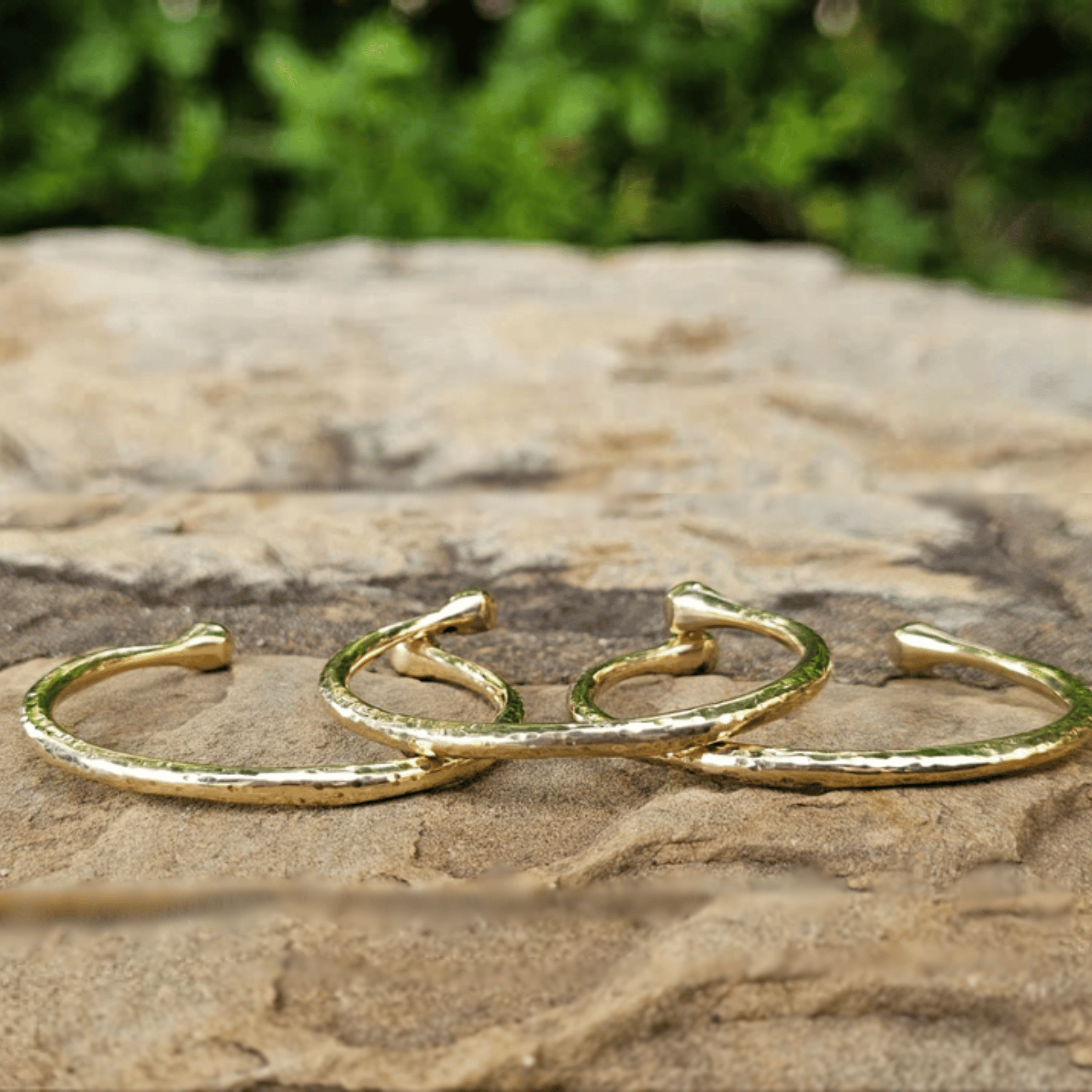 Three textured gold open cuff bracelets arranged on a natural stone surface with a blurred green outdoor background. Shop Kamawe