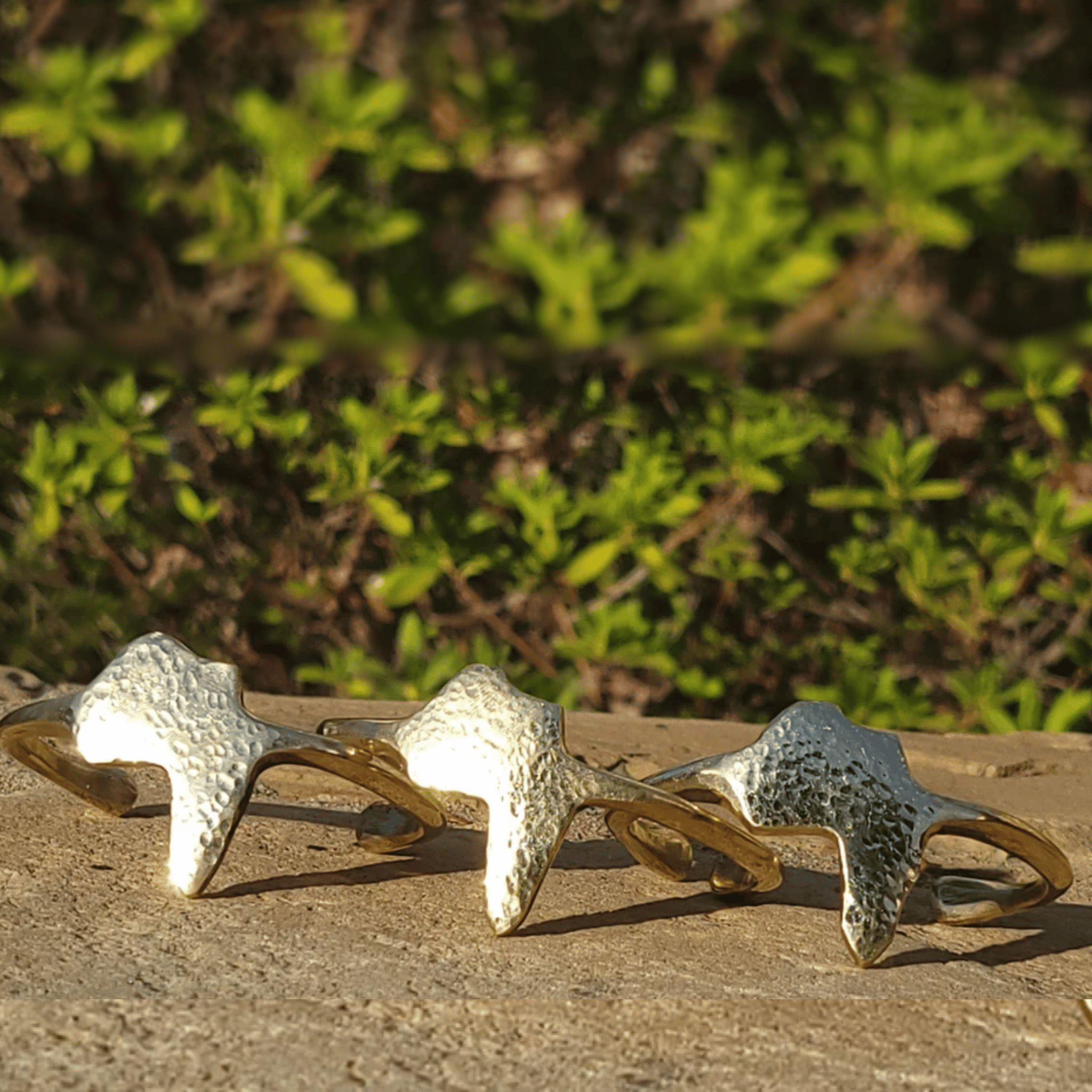 Three textured metallic rings with a hammered finish in silver and gold tones, arranged on a stone surface with a blurred green garden background. Shop Kamawe
