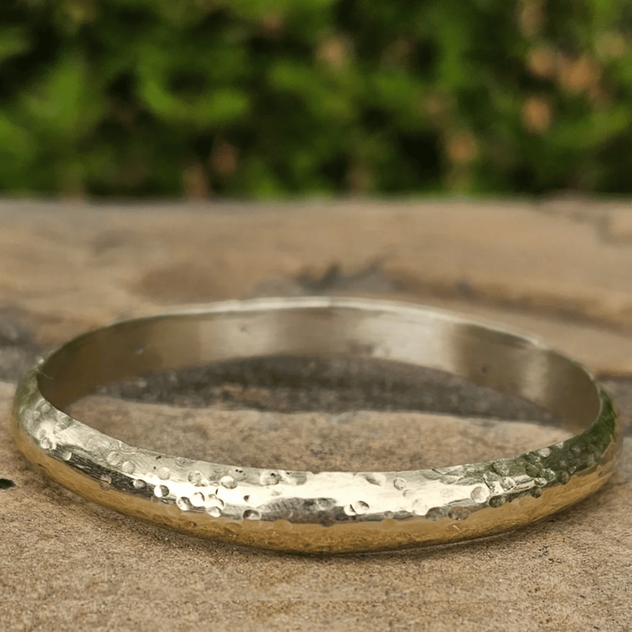 A hammered metal bangle bracelet in a silver-tone finish, resting on a textured stone surface with a blurred green outdoor background. Shop Kamawe