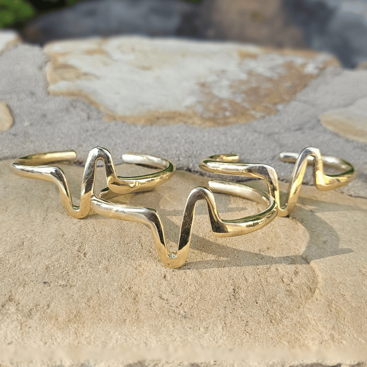 Two gold-toned open cuff bracelets with a wavy, flowing line design, displayed on a light beige stone surface. Shop Kamawe