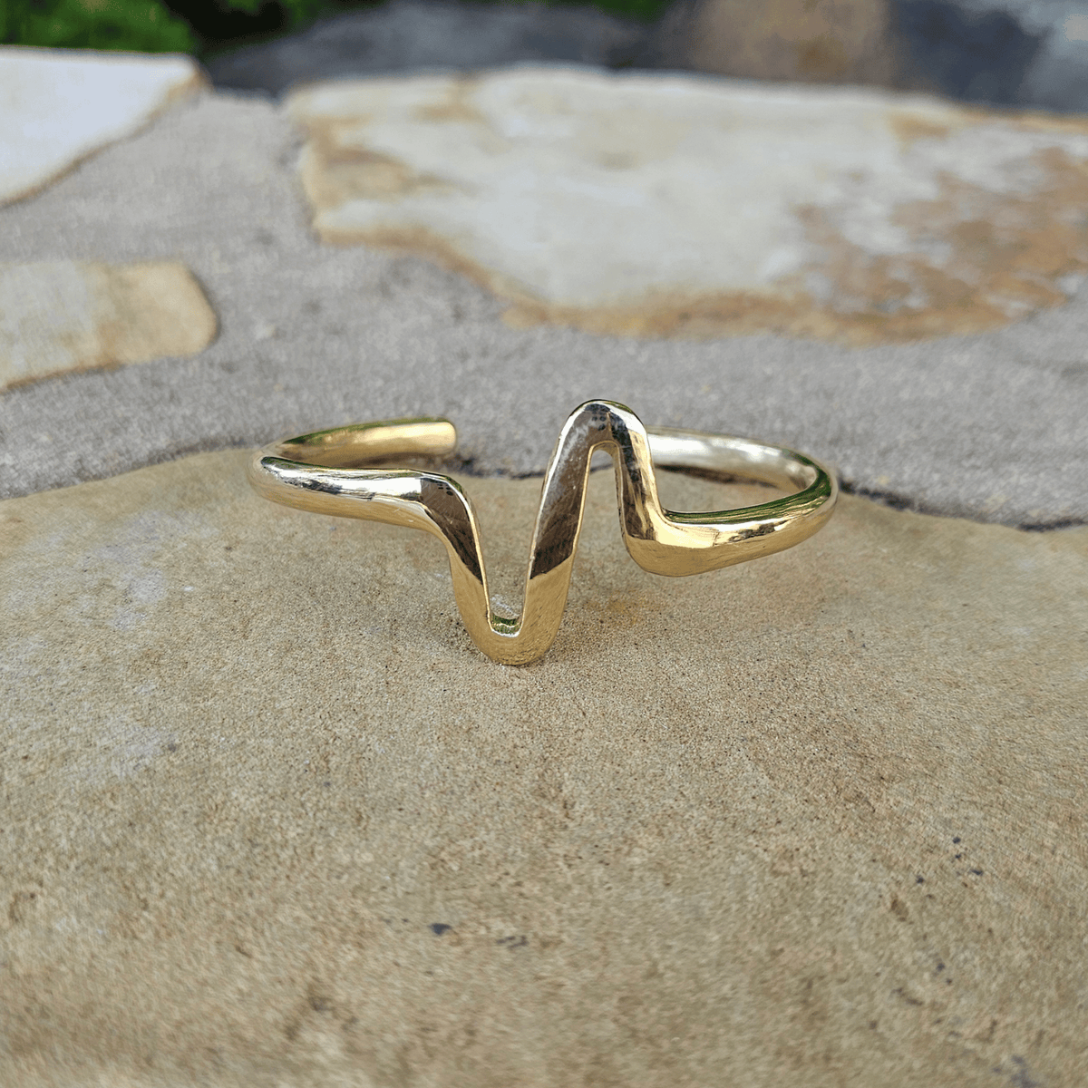 A gold-toned open cuff ring with a wavy, organic zigzag design, resting on a textured beige stone surface. Shop Kamawe