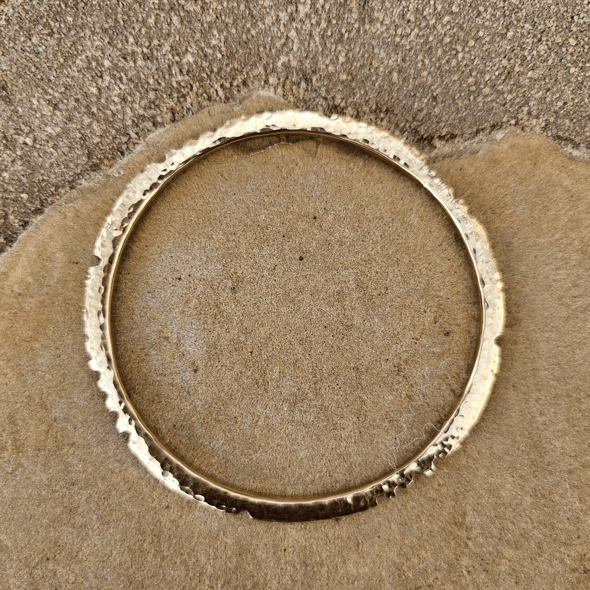 A hammered gold-toned metal bangle bracelet resting on a textured brown surface. Shop kamawe