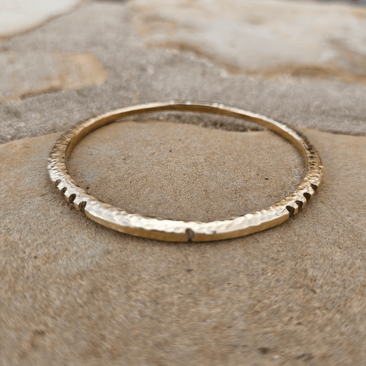 A hammered gold bangle bracelet resting on a textured sandy surface. Shop Kamawe
