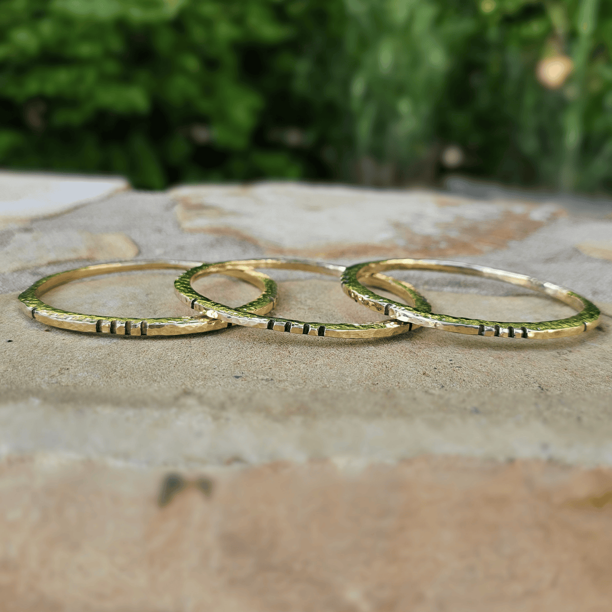 Three thin gold rings with small black horizontal stripes, arranged overlapping on a stone surface with a blurred green garden background. Shop Kamawe