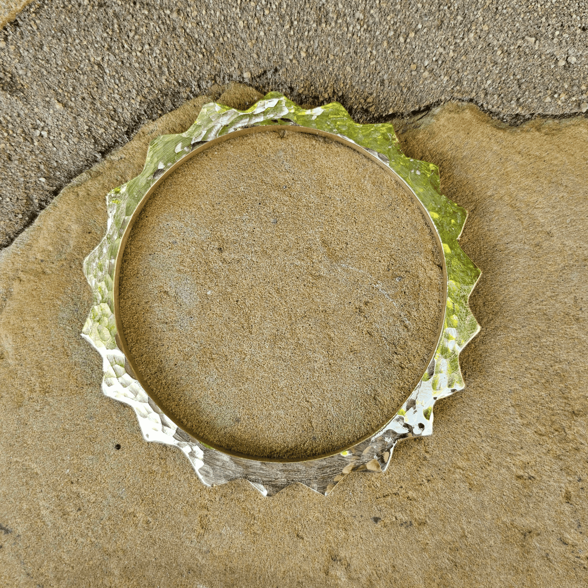 A hammered metal circular ring with a scalloped edge, shown from above on a textured tan stone surface. Shop Kamawe