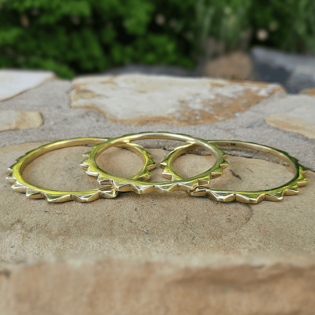 Three gold-toned spiked hoop earrings arranged on a stone surface outdoors, with blurred greenery in the background. Shop Kamawe