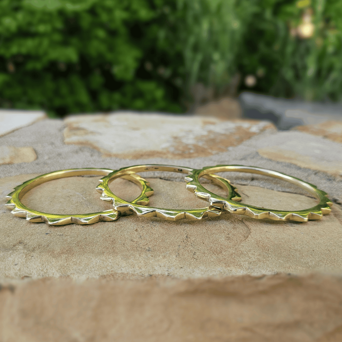 Three gold-toned spike-rimmed rings arranged overlapping on a stone surface with a blurred green outdoor background. Shop Kamawe