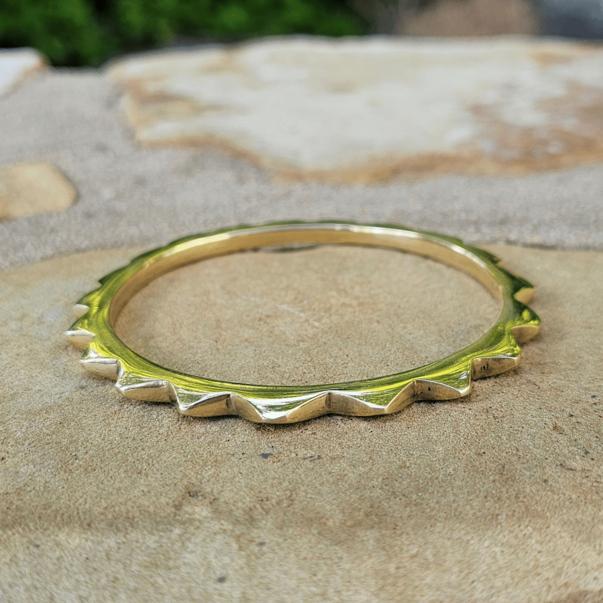 A gold-toned metal bangle bracelet with a textured, spiked outer edge, resting on a natural stone surface with a blurred outdoor background. Shop Kamawe
