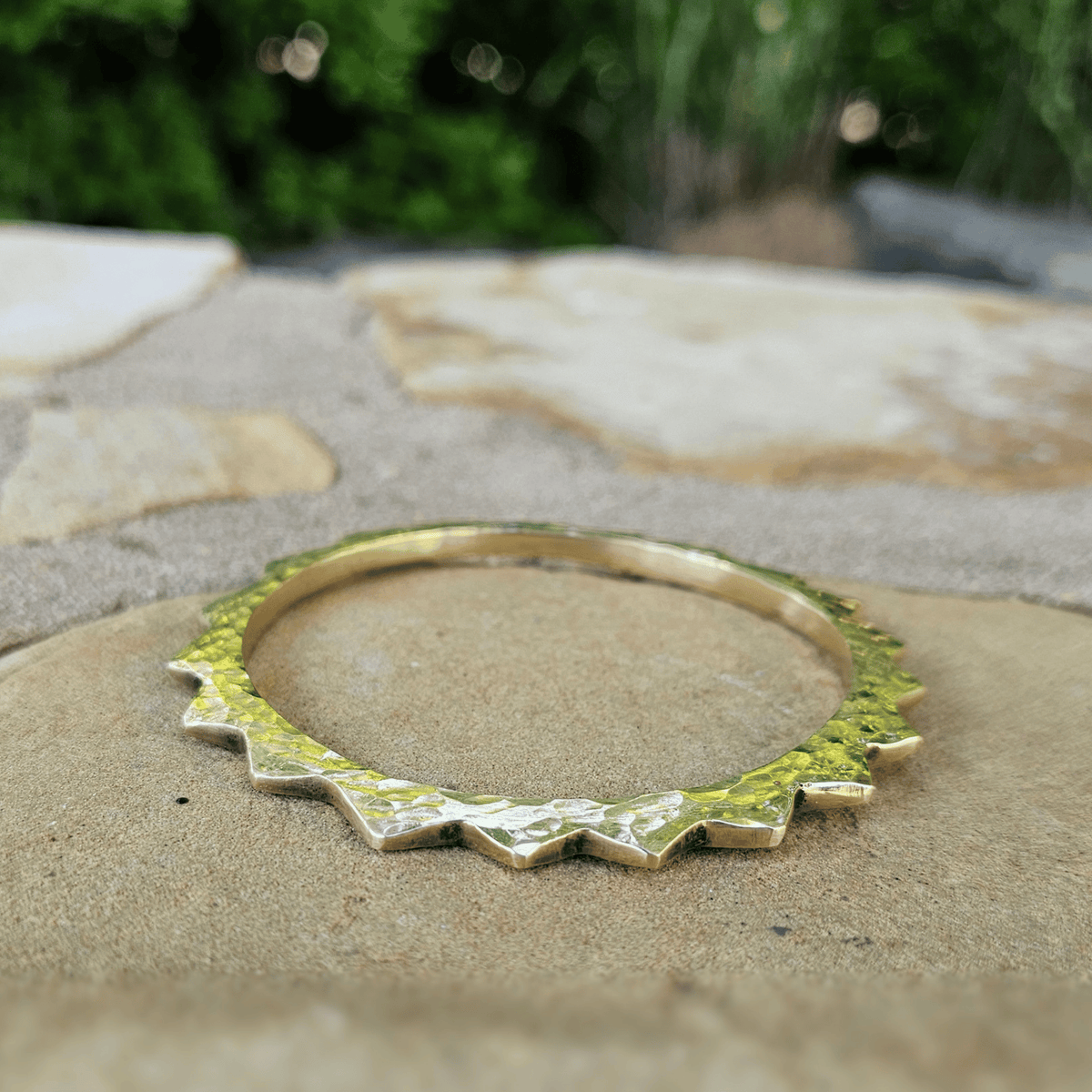 A hammered gold bangle bracelet with a jagged, scalloped edge, resting on a stone patio with a blurred green garden background. Shop Kamawe
