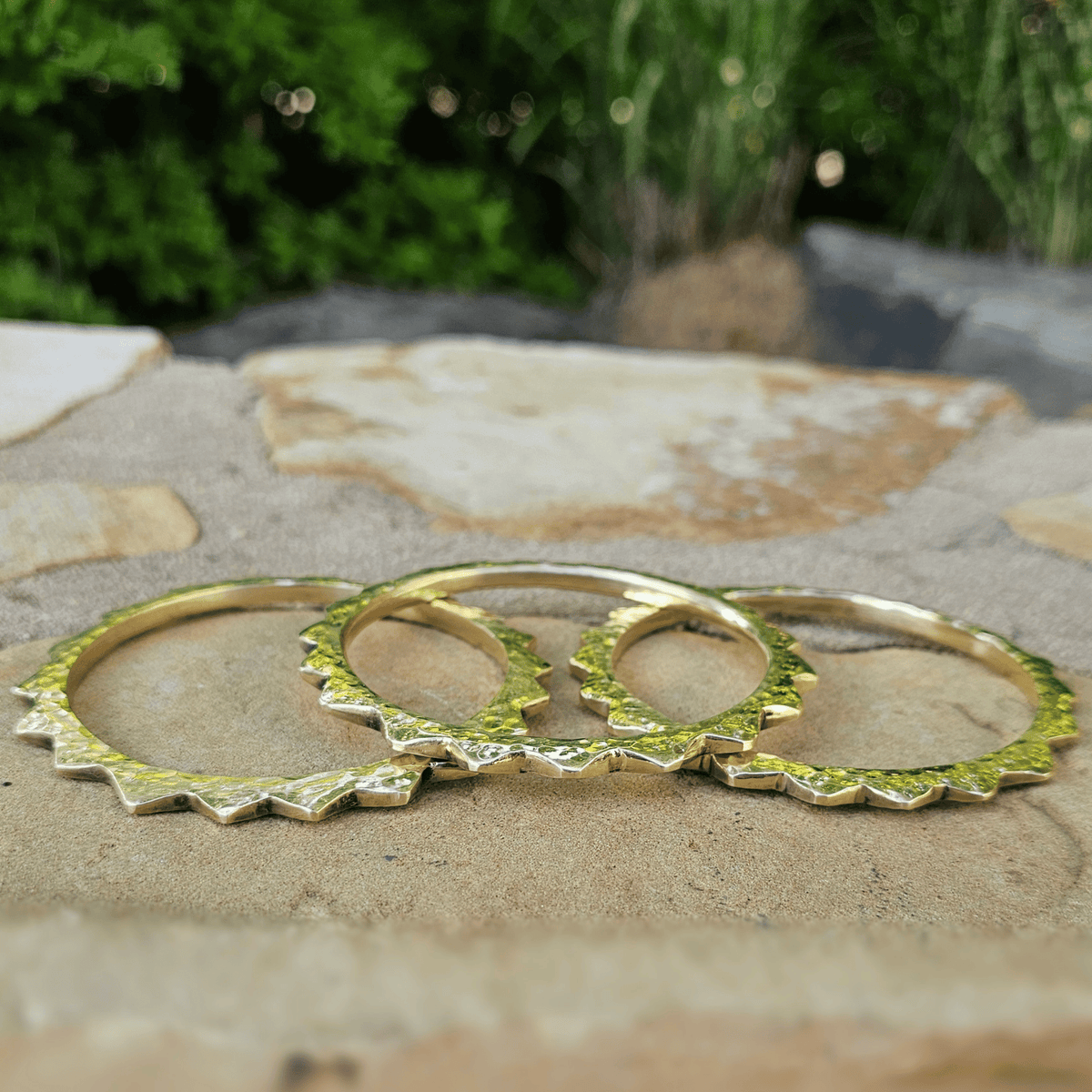 Three gold-toned rings with textured, jagged scalloped edges arranged on a stone surface with a blurred green outdoor background. Shop Kamawe