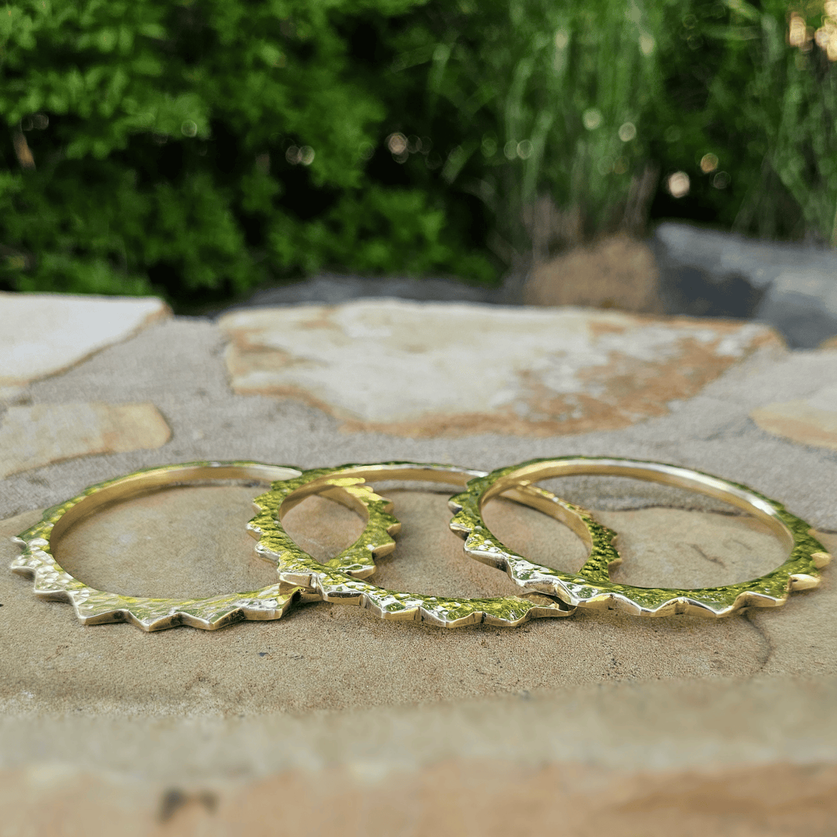 Three gold-toned textured bangles with jagged edges, displayed on a stone surface with a blurred green garden background. Shop Kamawe
