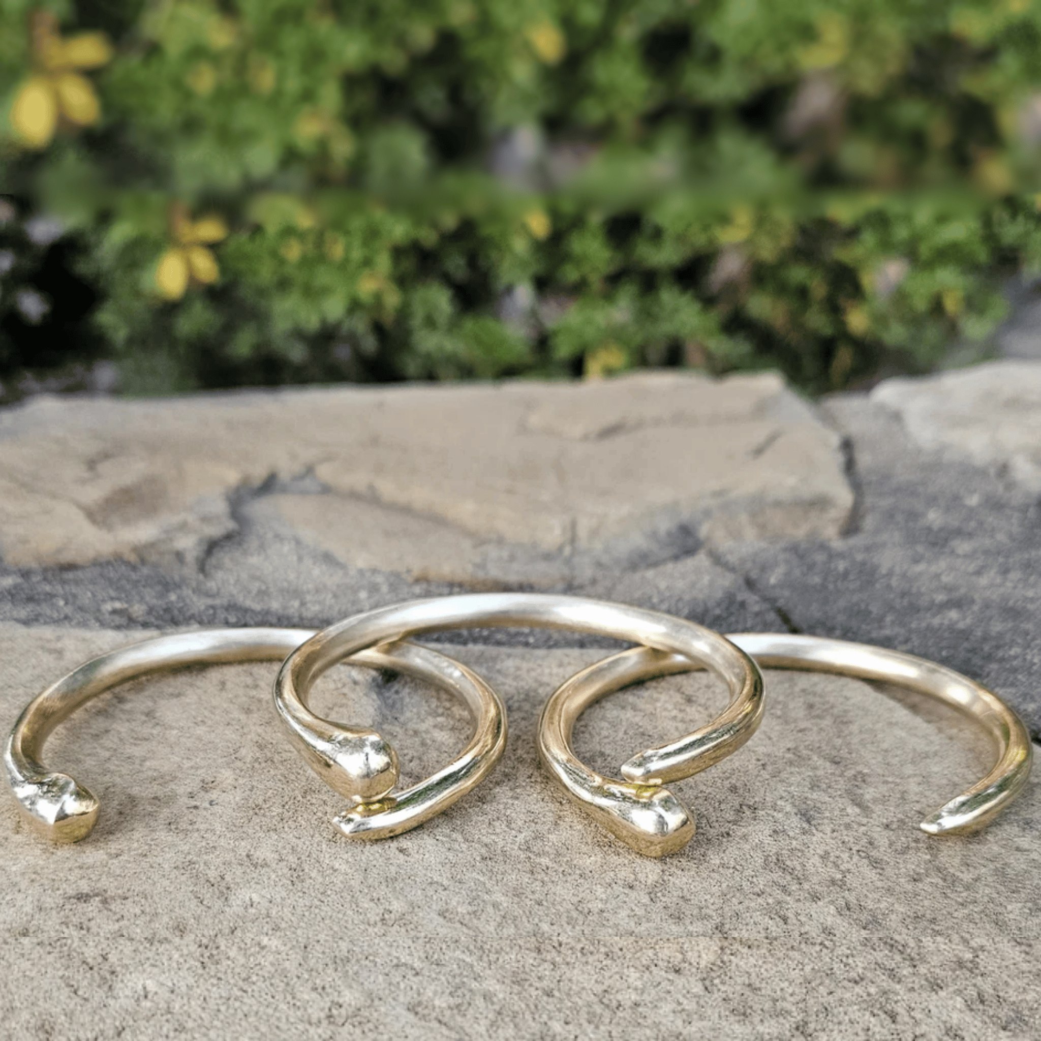 Three gold-toned open cuff bracelets with textured, organic ends resting on a stone surface with a blurred green garden background. Shop Kamawe
