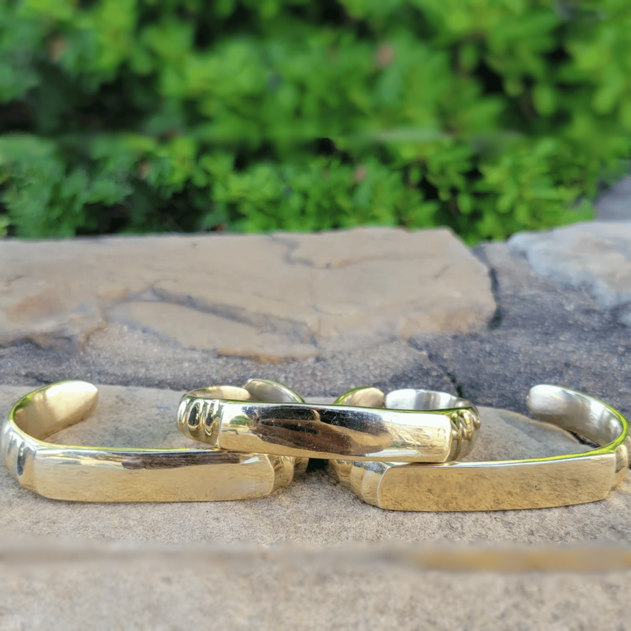 Three polished gold-toned metal cuff bracelets resting on a stone surface with a blurred green foliage background. Shop Kamawe