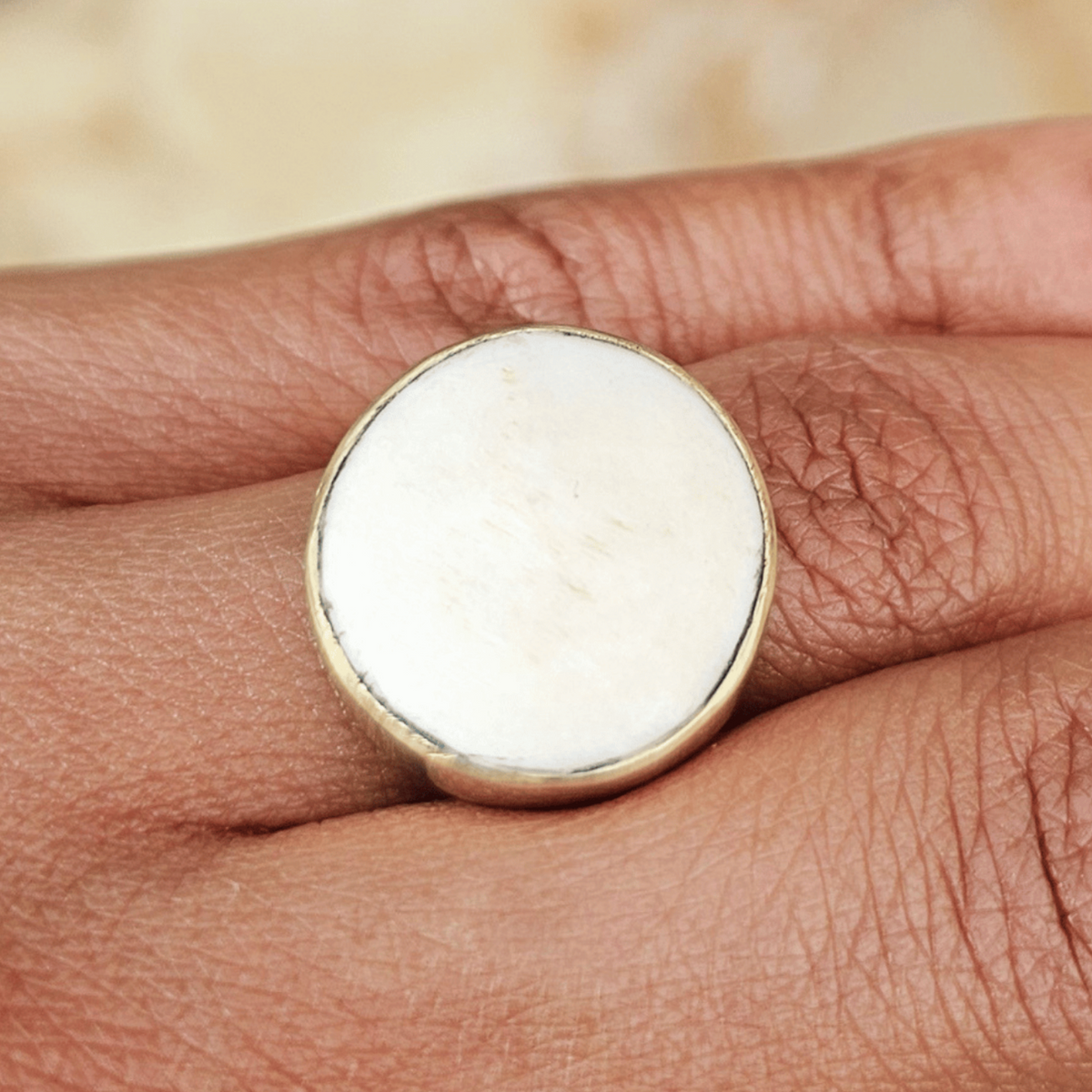 A close-up of a hand wearing a large round signet ring with a white stone or inlay set in a gold-toned bezel, against a softly blurred neutral background. Shop Kamawe