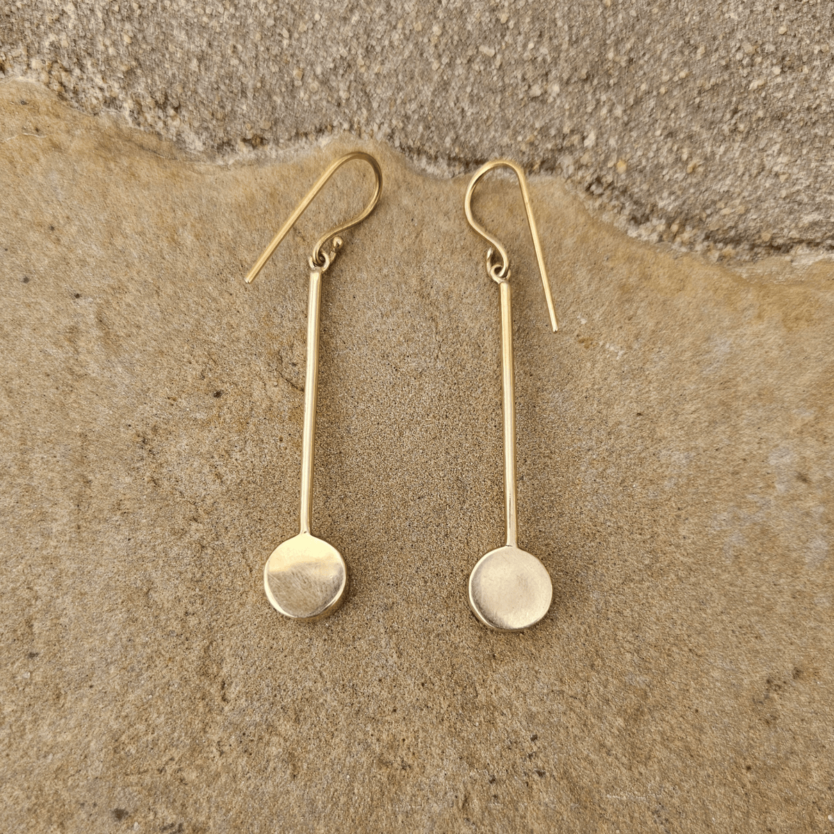 A pair of simple gold drop earrings with flat circular disc pendants and fishhook backs, displayed on a textured beige stone surface. Shop Kamawe