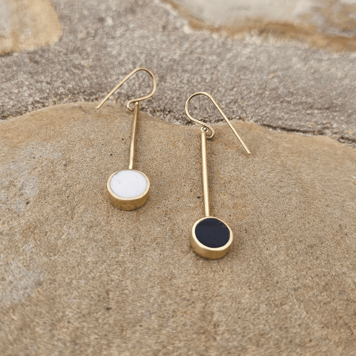 A pair of gold drop earrings with circular disc charms, one white and one dark navy blue, resting on a textured sandy surface. Shop Kamawe