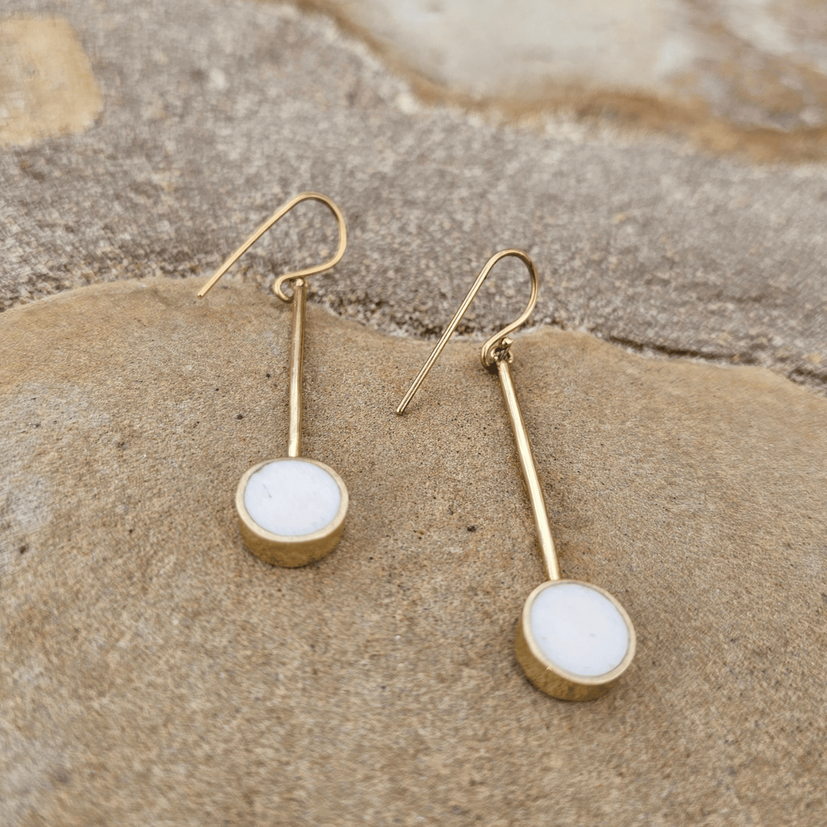 A pair of gold drop earrings with round white cabochon stones, displayed on a textured beige rock. Shop Kamawe