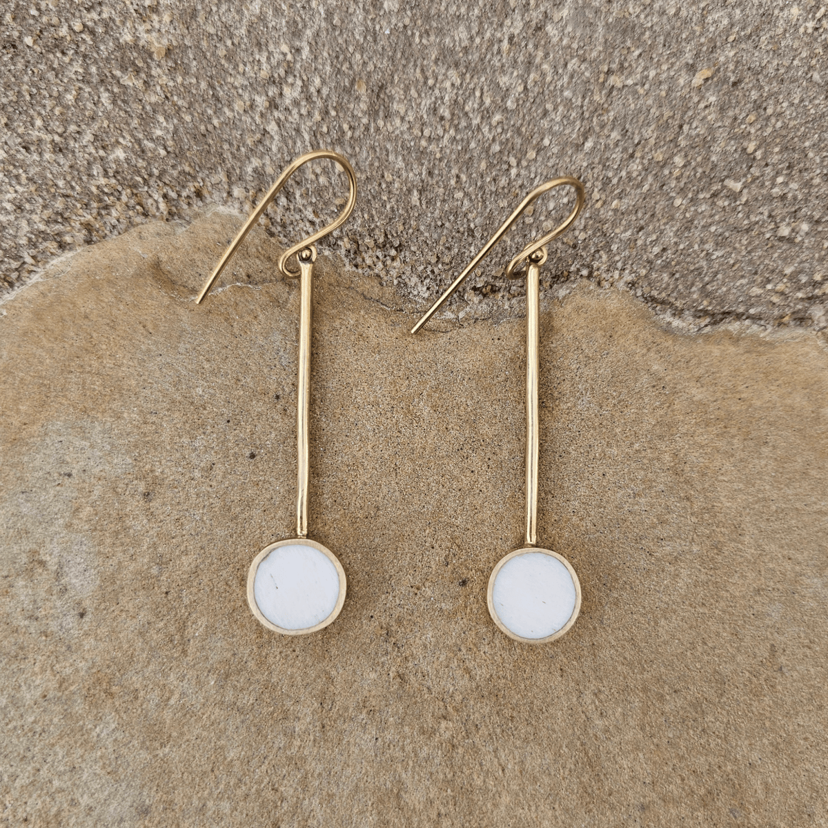 A pair of gold dangle earrings with round white stone bezel settings, displayed on a textured tan stone surface. Shop Kamawe