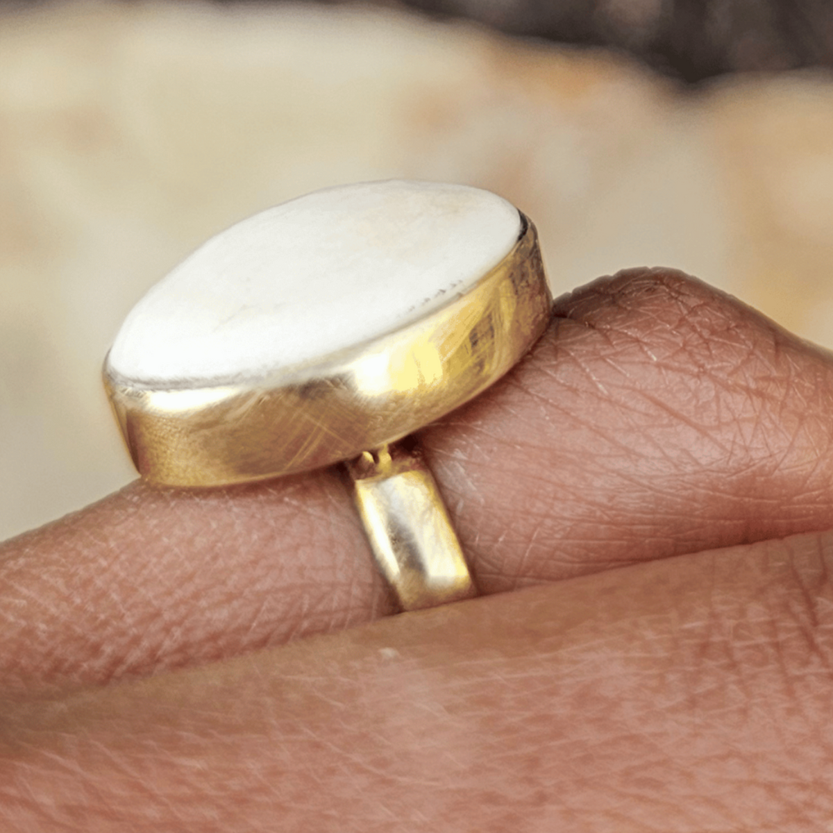 A gold ring with a large, polished white oval stone worn on a person's finger, set against a blurred natural background of earth and light-colored stones. Shop Kamawe
