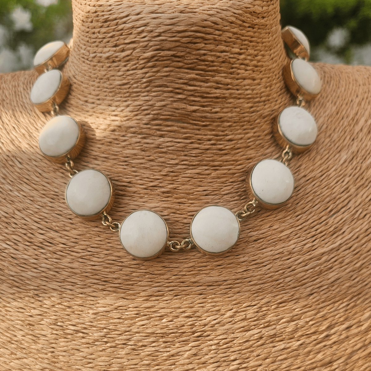 A statement necklace with large round off-white cabochon stones set in gold-tone metal, displayed on a textured tan woven jewelry bust. Shop Kamawe