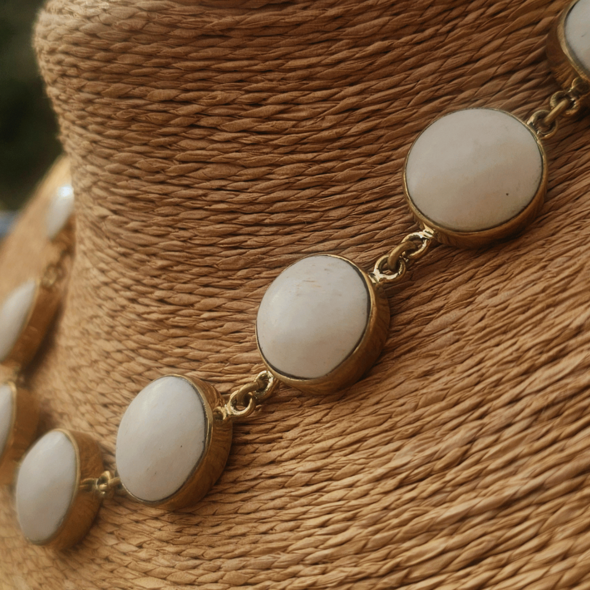 A gold-tone chain necklace with oval white stone accents, displayed on a woven tan mannequin bust with an outdoor blurred background. Shop Kamawe
